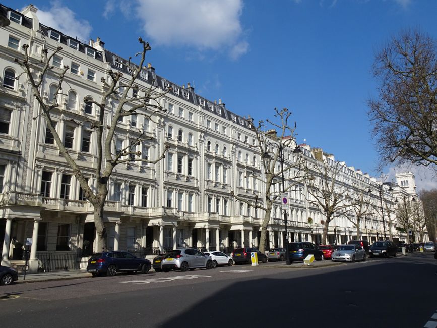 A row of elegant, white Victorian-style terraced buildings with decorative cornices and large sash windows, situated along a tree-lined street in St Johns Wood. Several leafless trees with pruned branches line the pavement, which is wide and clean. Parked cars of various makes and colours are aligned along the curb, with some yellow and black bollards at the street edge. The sky is bright blue with a few scattered clouds, and the scene is illuminated by natural daylight. This image illustrates a typical residential street within the St. John's Wood area, relevant for house removals and furniture transport services offered by Movers St. John's Wood, including the process of home relocation and loading activities on the pavement.