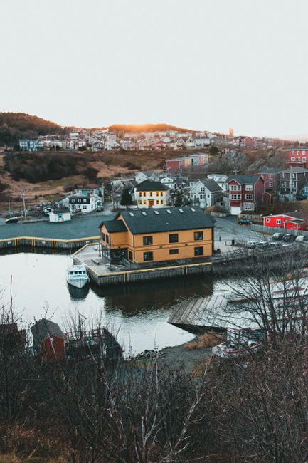 A view of a coastal residential area during sunset, featuring a small house with a yellow-brown exterior and black roof positioned on a narrow pier extending over calm water. In front of the house, there is a white boat docked at the floating platform. Surrounding the water are several other houses and buildings with varied architecture, including some with red and blue facades, situated on a gently sloping hillside with trees and open spaces. The background includes a distant town with numerous houses and structures, and the sky displays warm orange hues as the sun sets behind the hilltops. This scene depicts a serene home environment, which could be part of a property undergoing moving or relocation services by a company such as Movers St. John's Wood, evident from the context of house removals and moving logistics.